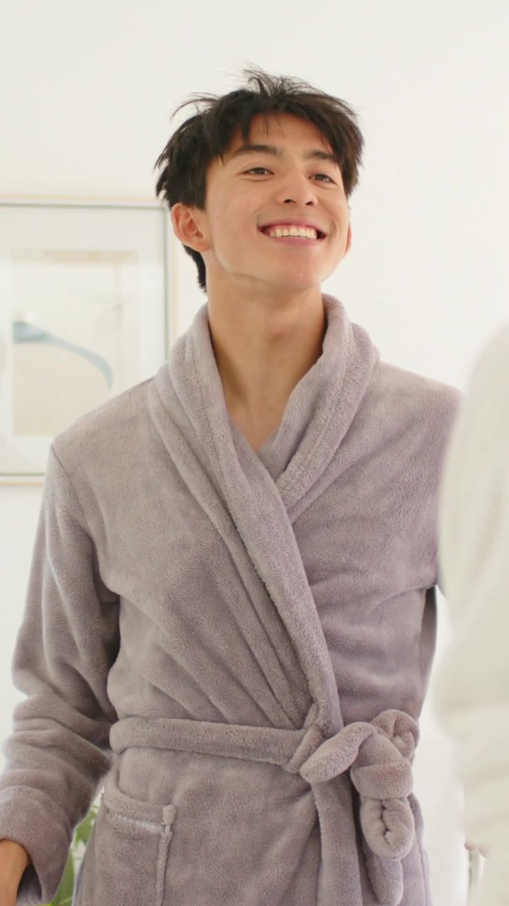 Vertical video: Grabbing rinse cup, man in gray robe checking teeth at home mirror, partner joining