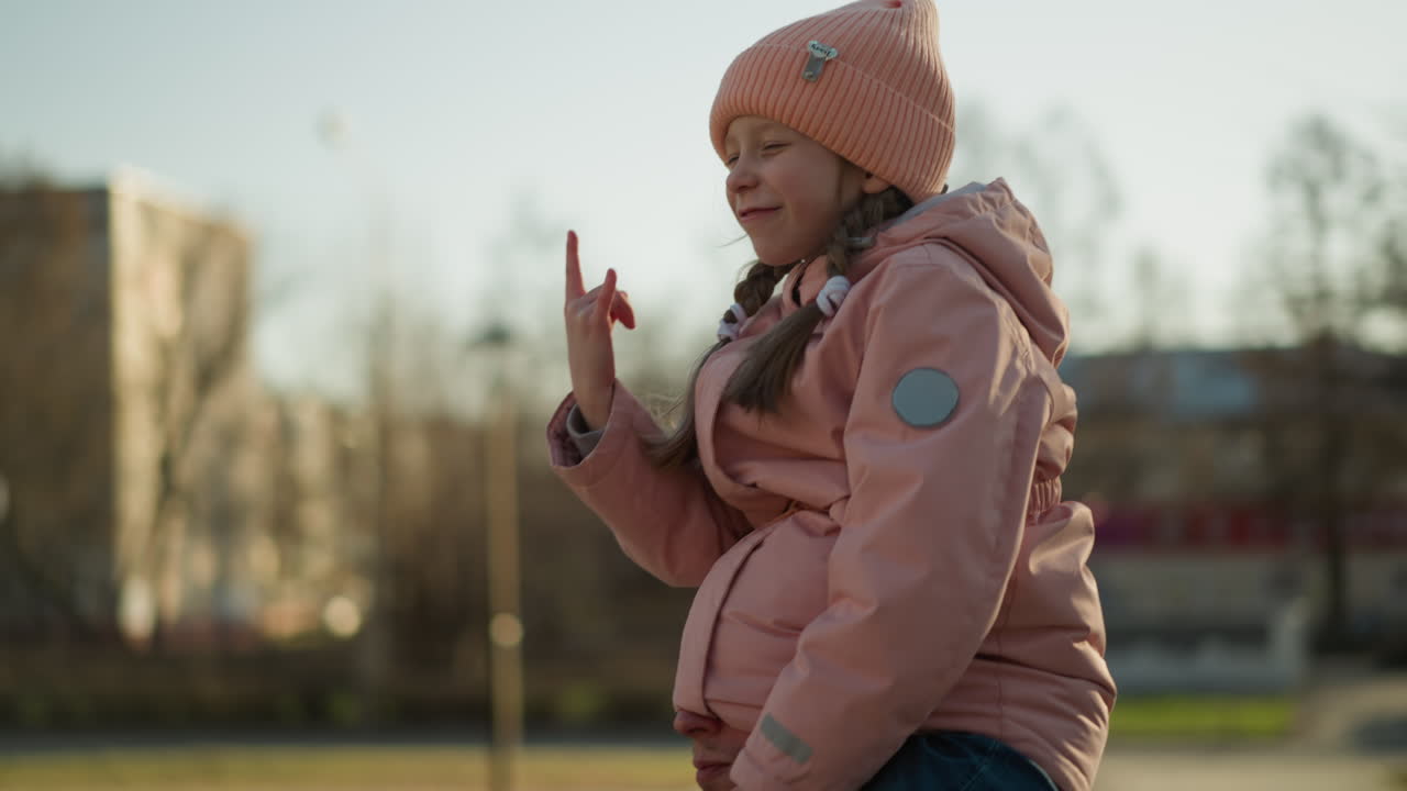 핑크색 모자와 재을 입은 어린 소녀는 아버지의 목에 기게 앉아 있으며, 그의 머리의 절반은 재게 그녀의 재에 혀 있으며, 그녀는 장난스러운 손짓을 하고 미소 짓고 있습니다.