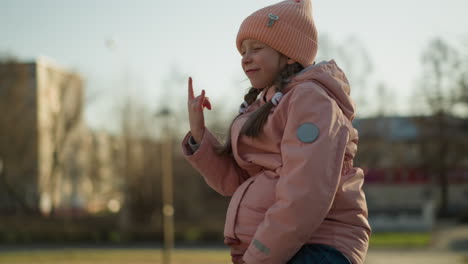 una niña con una gorra rosa y una chaqueta se sienta alegremente en el cuello de su padre, con la mitad de su cabeza bien metida en su chaqueta mientras hace una señal de mano lúdica y sonríe