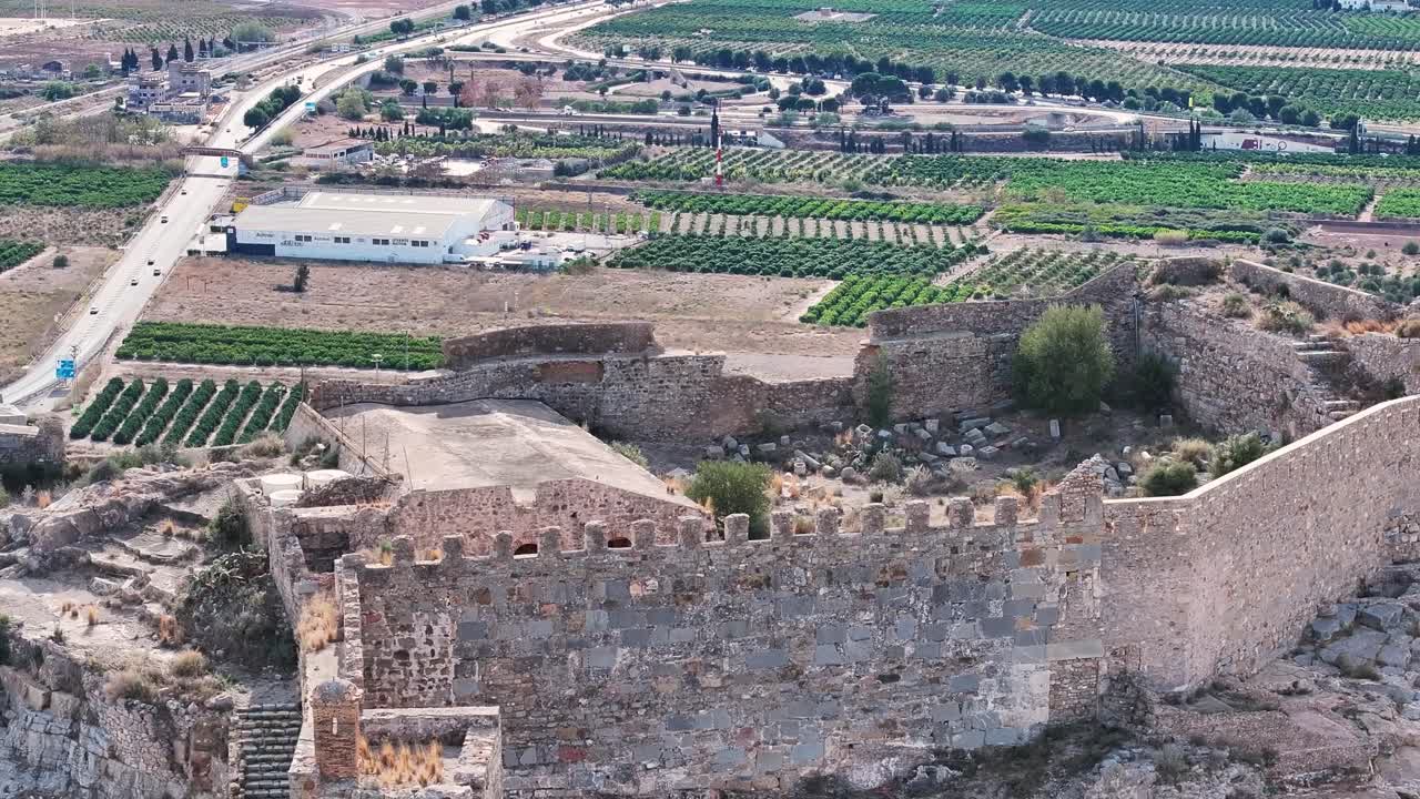 vuelo donde vemos una parte del castillo donde guardan restos arqueológicos y en el fondo hay cultivos de naranjos y algunas carreteras con vehículos circulando vemos un efecto de paralaje