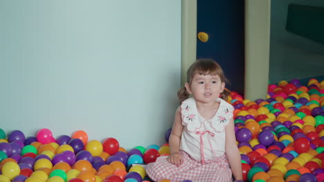 cheerful excited toddler girl in ball pit throws a ball in a playroom filled with colorful plastic balls