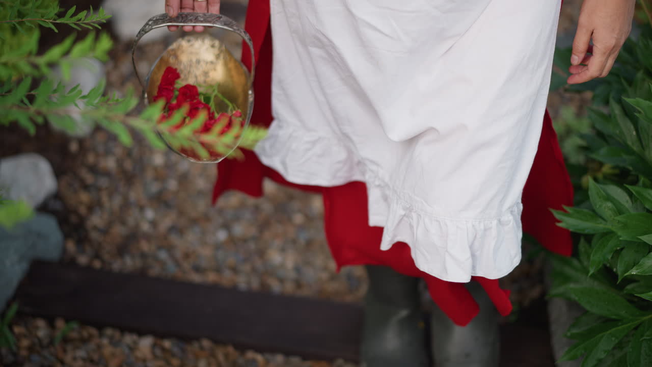 Red dress white apron holding basket, downward camera view on wooden steps and pebble path, rubber boots stepping over boards, florist delivering red carnations, homegrown market routine in warm