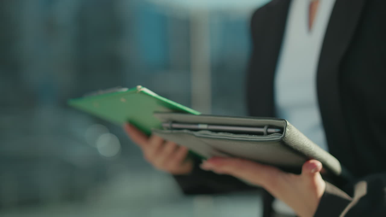 Close up of professional individual holding folder and documents, cross checking details while standing outdoors with glass building in background, focused on reviewing information