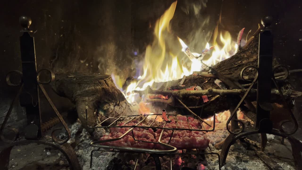 Salsicce in cottura sulla griglia del camino con fuoco acceso sullo sfondo, primo piano.