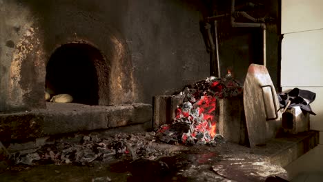 Alguien-Poniendo-Un-Poco-De-Pan-En-Un-Viejo-Horno-De-Leña-Junto-A-Una-Chimenea-Llena-De-Carbón-Rojo-Y-Caliente