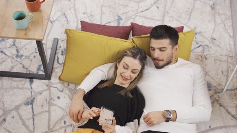 una pareja joven sentada en casa usando un teléfono inteligente, comprando productos en línea, planeando vacaciones, reservando un hotel.