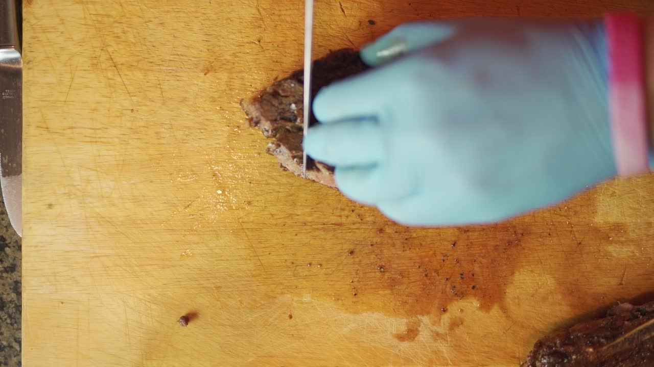 Vista dall'alto, filmato al rallentatore, di un uomo che indossa guanti mentre affetta striscioline di bistecca cotta e succosa su un tagliere di legno con un grande coltello da chef