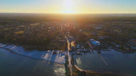 aerial view of a small coastal town and marina at sunset, with soft golden light illuminating buildings, boats, docks, and surrounding forest landscape