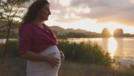 pregnant woman expectant mother walking alone by lake at sunset, attractive caucasian casual dressed mom stroking caressing touching her belly with hands smiling, childbearing medium portrait shot