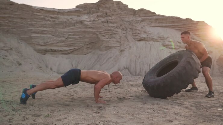 Two muscular open-chested athletes train in active mode on the beach doing push-UPS and pushing a huge wheel against a sandy mountain at sunset