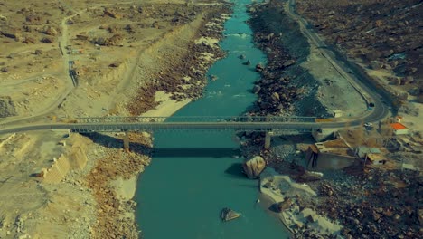 Aerial-view-capturing-a-bridge-spanning-a-blue-river-amidst-rugged,-rocky-terrain