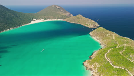 A-drone-ascends-capturing-a-breathtaking-view-of-Arraial-do-Cabo's-stunning-blue-turquoise-bay-with-a-picturesque-island-shoreline-and-majestic-mountains-in-the-background