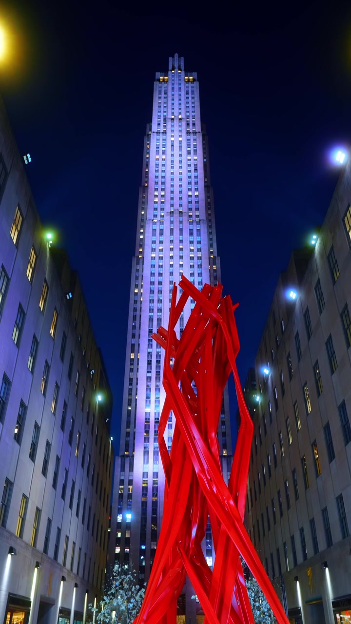 Low angle view of Rockefeller Center with red sculpture. The 30 Rockefeller Plaza skyscraper illuminated at night with a red abstract sculpture in the foreground.