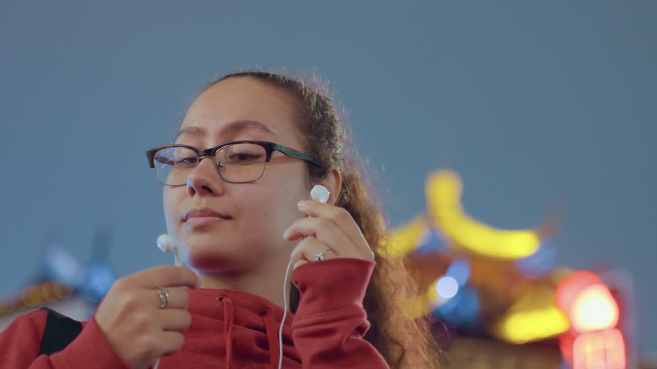 Junge Frau im roten Kapuzenpullover justiert Kopfhörer neben einer beleuchteten Tempelfassade, setzt Ohrhörer mit konzentriertem Ausdruck ein, Vlogger- und Reiseenergie, Lichter des städtischen Nachtmarkts und Bokeh, selbstbewusst.