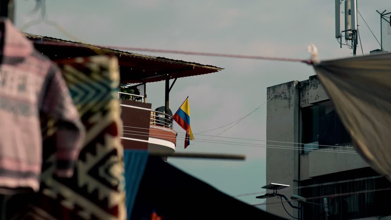 Immagine della bandiera ecuadoriana con lo stemma che sventola su un balcone moderno, incorniciata da tessuti tradizionali andini e architettura urbana