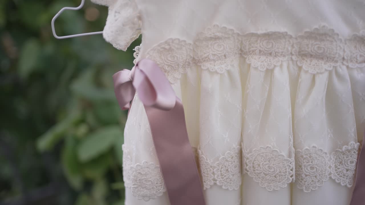 vestido de bebé delicado con detalles de encaje y un lazo rosa, colgado en una percha al aire libre, perfecto para una ocasión especial