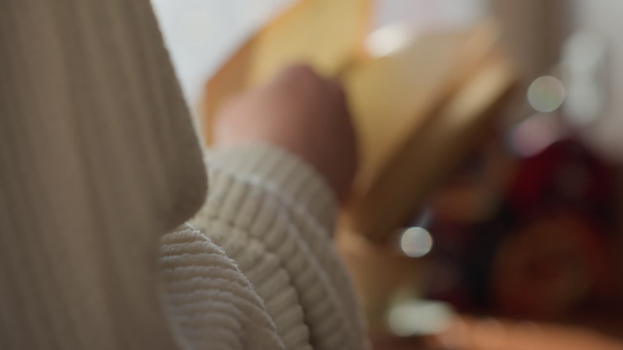 Close up of lady hand flipping page of open book in soft daylight near window, cozy setting includes blurred mug on wooden table and lush green plant in background