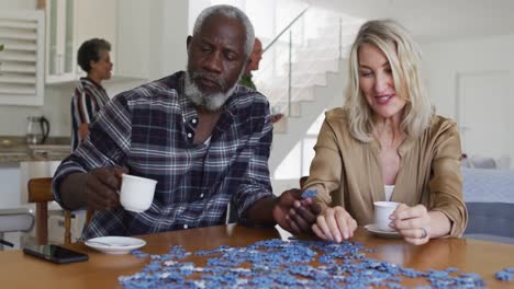 homem idoso afro-americano e mulher idosa caucasiana sentados à mesa fazendo quebra-cabeças bebendo chá