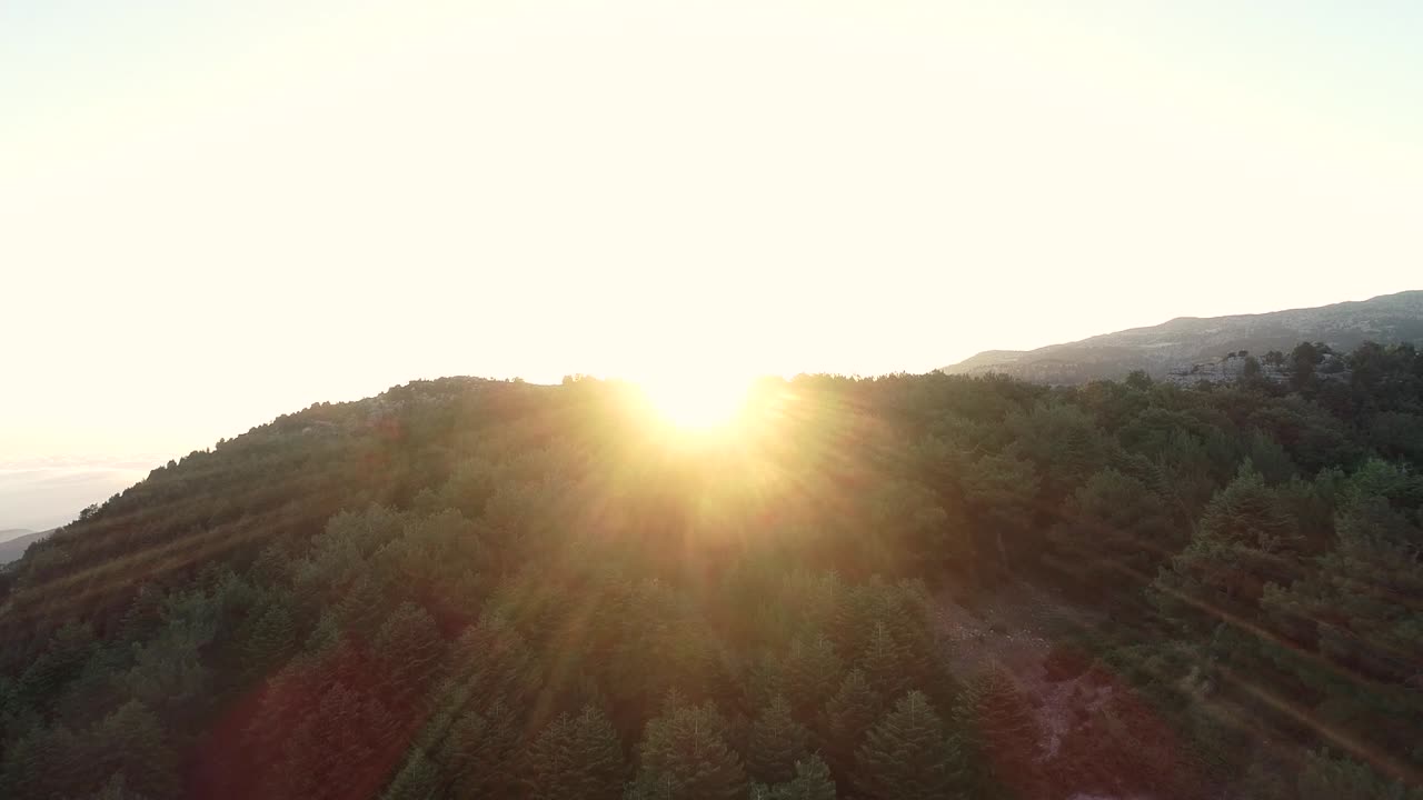 sunrise appears with lens flare in the top of the mountain and a beautiful scenery. Aerial shot flying over the trees and revealing the sunrise behind the mountain.