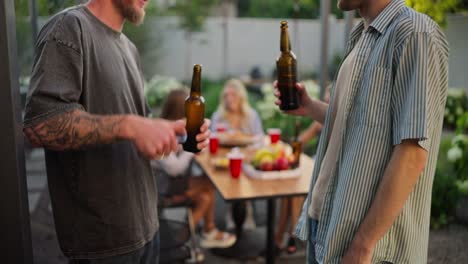 vista lateral de una feliz chica rubia y sus dos amigas hablando mientras los chicos tocan vasos con botellas marrones de cerveza y se divierten en el patio de una casa de campo