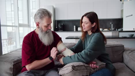 An-elderly-man-with-gray-hair-and-a-lush-beard-in-a-red-T-shirt-helps-his-adult-brunette-daughter-in-a-Green-jacket-bandage-his-hand-with-bandages-while-sitting-on-the-sofa-in-a-modern-apartment