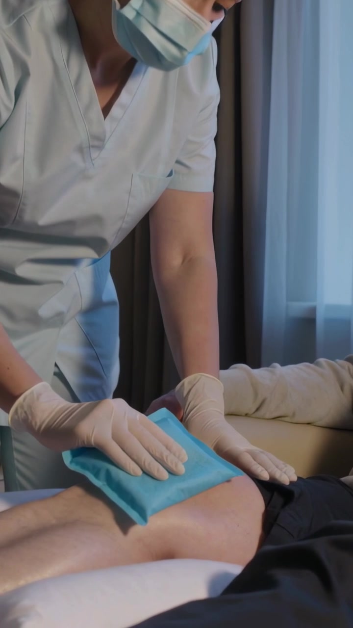 Video vertical: Enfermera quirúrgica colocando compresa fría en la rodilla sobre una camilla de examen para aliviar el dolor.
