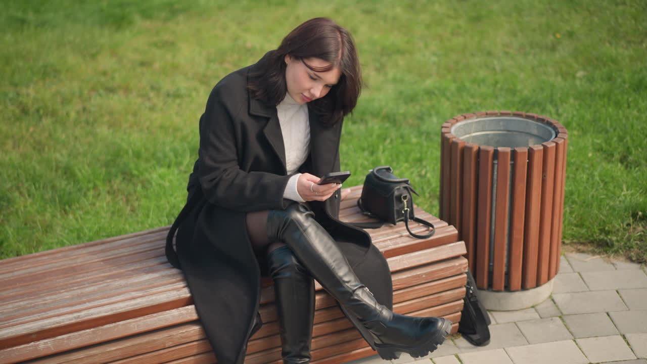 молодая девушка в черном пальто и белом свитере сидит на скамейке в парке, сосредотачиваясь на смартфоне с сумкой поблизости, расслабленная атмосфера на открытом воздухе, в окружении зелени в городском парке