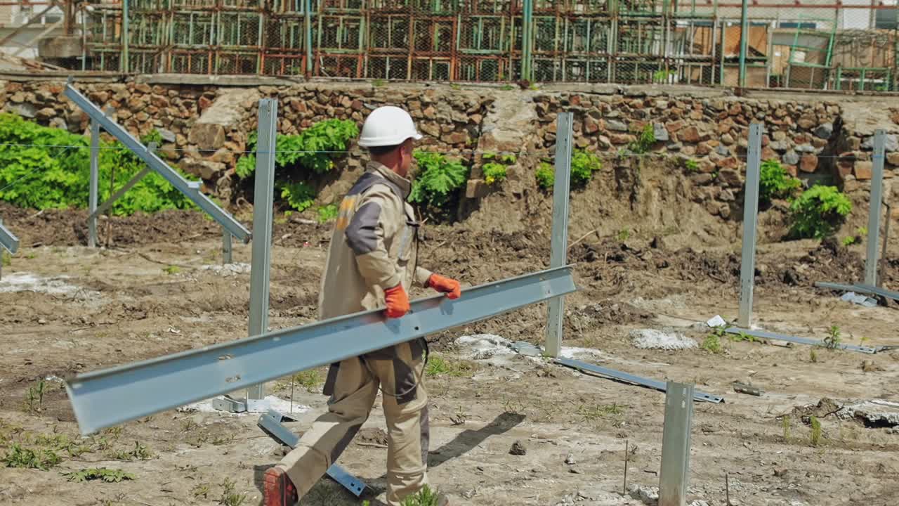 Construction site of a solar farm outdoors. Worker in safety helmet carries metal frame on the field.