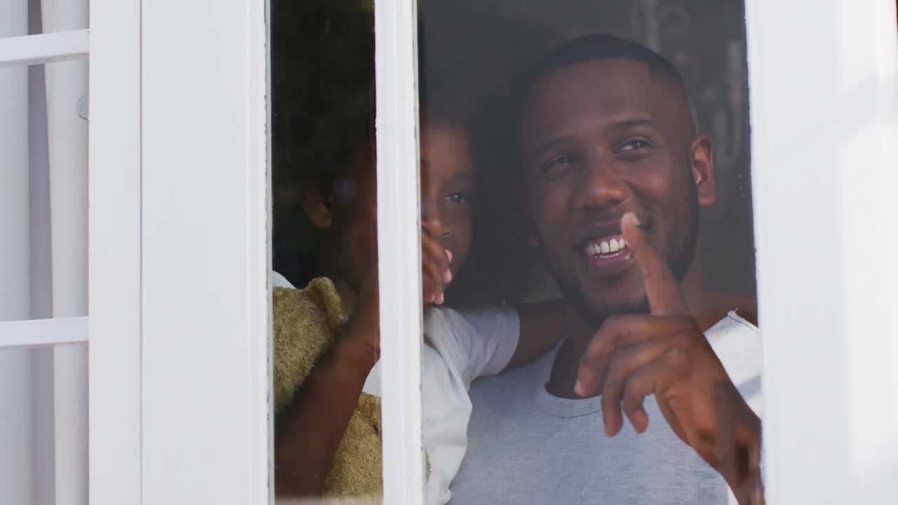 African american daughter holding teddy bear looking through window with her father