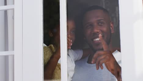 African american daughter holding teddy bear looking through window with her father