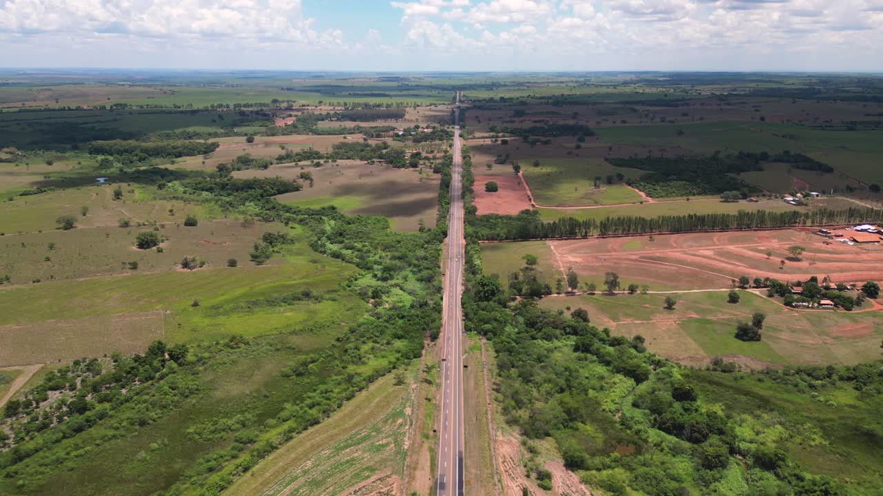 Foto aerea dall'alto di una lunga e rettilinea autostrada che divide ampi campi e terreni agricoli rurali nell'entroterra dello Stato di San Paolo, in Brasile. Cieli limpidi e ampi spazi aperti suggeriscono libertà e viaggi.
