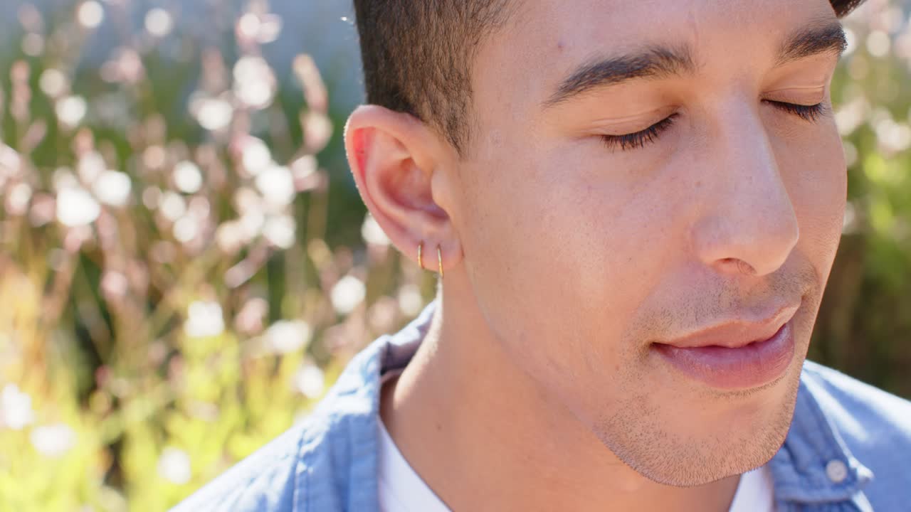 Un uomo afroamericano si rilassa, girandosi mentre la telecamera si sposta nel giardino, indossando una camicia blu e doppi orecchini a cerchio.