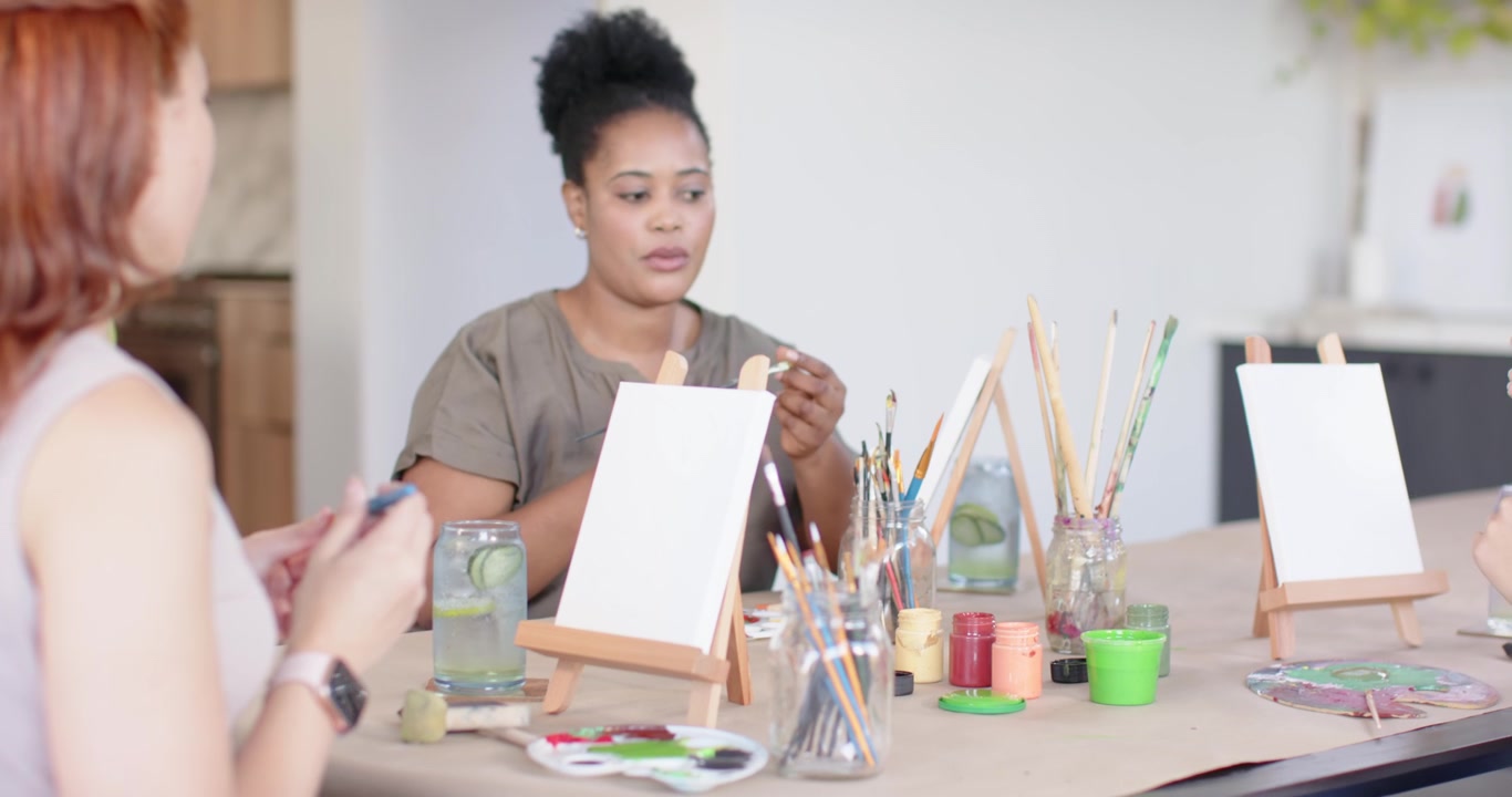 Diverse women checking brushes, dipping brush, readying canvas while camera pulling back at studio