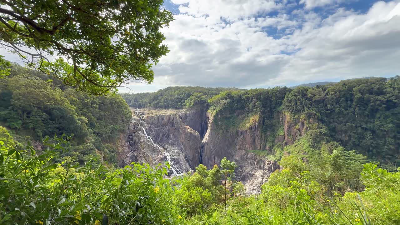 Train moves along forested cliff above dramatic waterfall, lush greenery, and bright daylight landscape