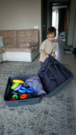 a big opened suitcase filled with toy pyramid parts lies on the floor. little baby boy runs into the room, throws the car into the suitcase and closes it. vertical video.
