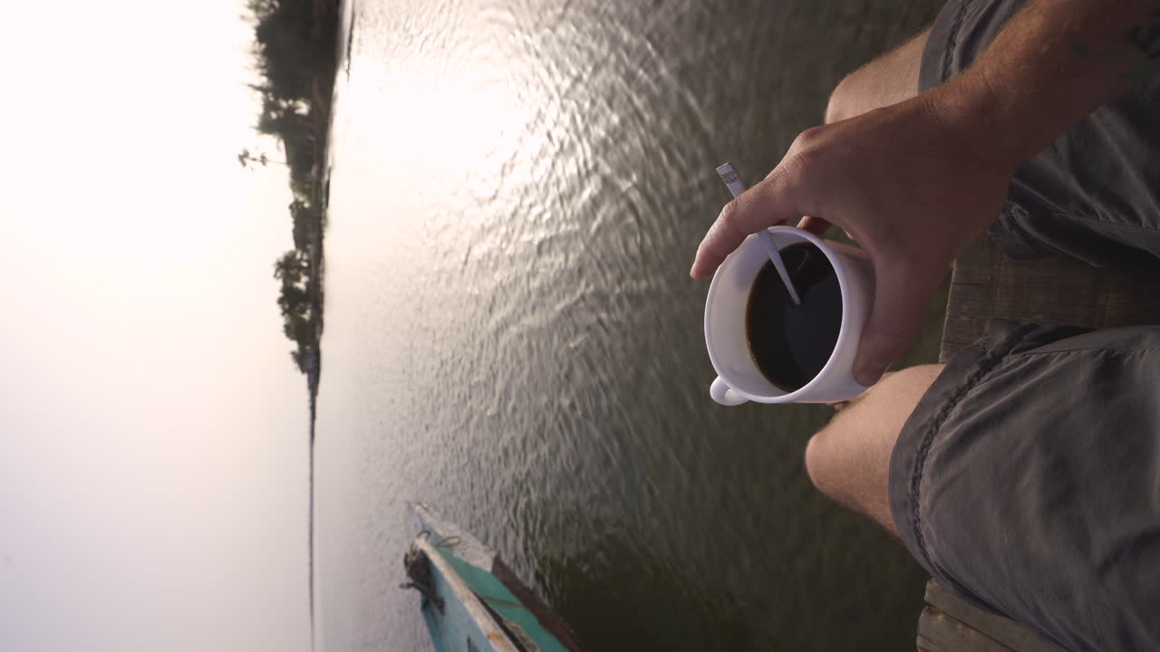 vertical de un joven aventurero explorador caucásico bebiendo una taza de café negro mientras se sienta en la orilla del río en senegal áfrica durante la puesta de sol