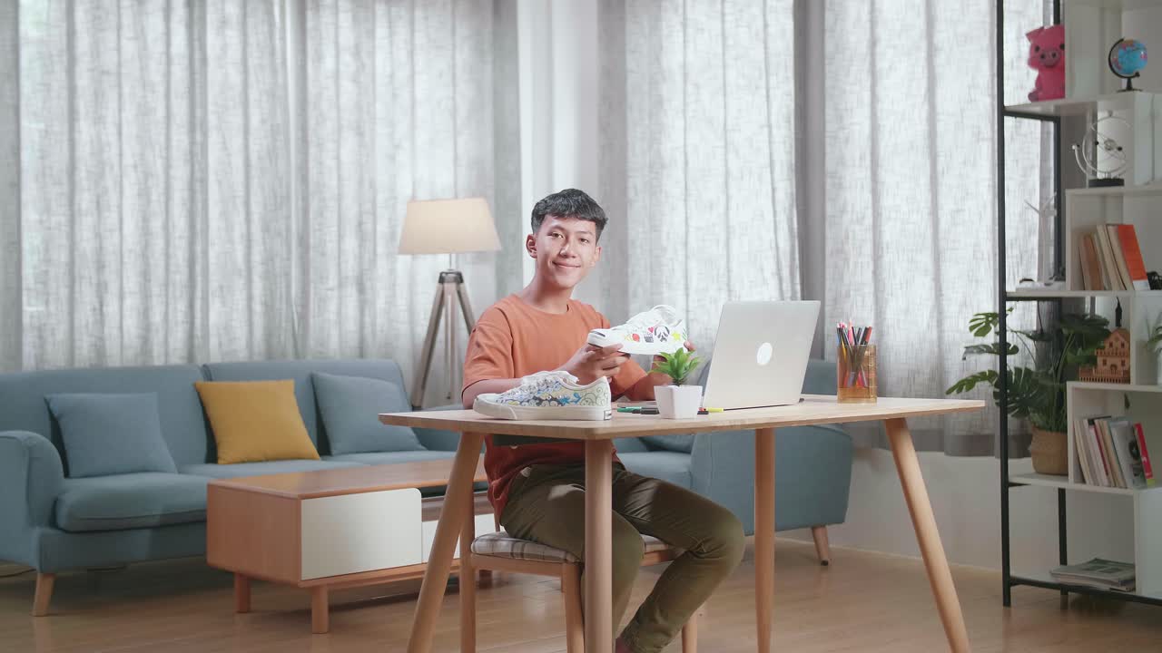 아시아 소년 신발 디자이너가 집에서 노트북을 사용하면서 카메라에 미소 짓고 있습니다.