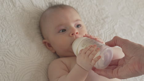 Baby-sucking-on-milk-bottle,-newborn-feeding-time-with-mommy-in-white-bright-bedroom