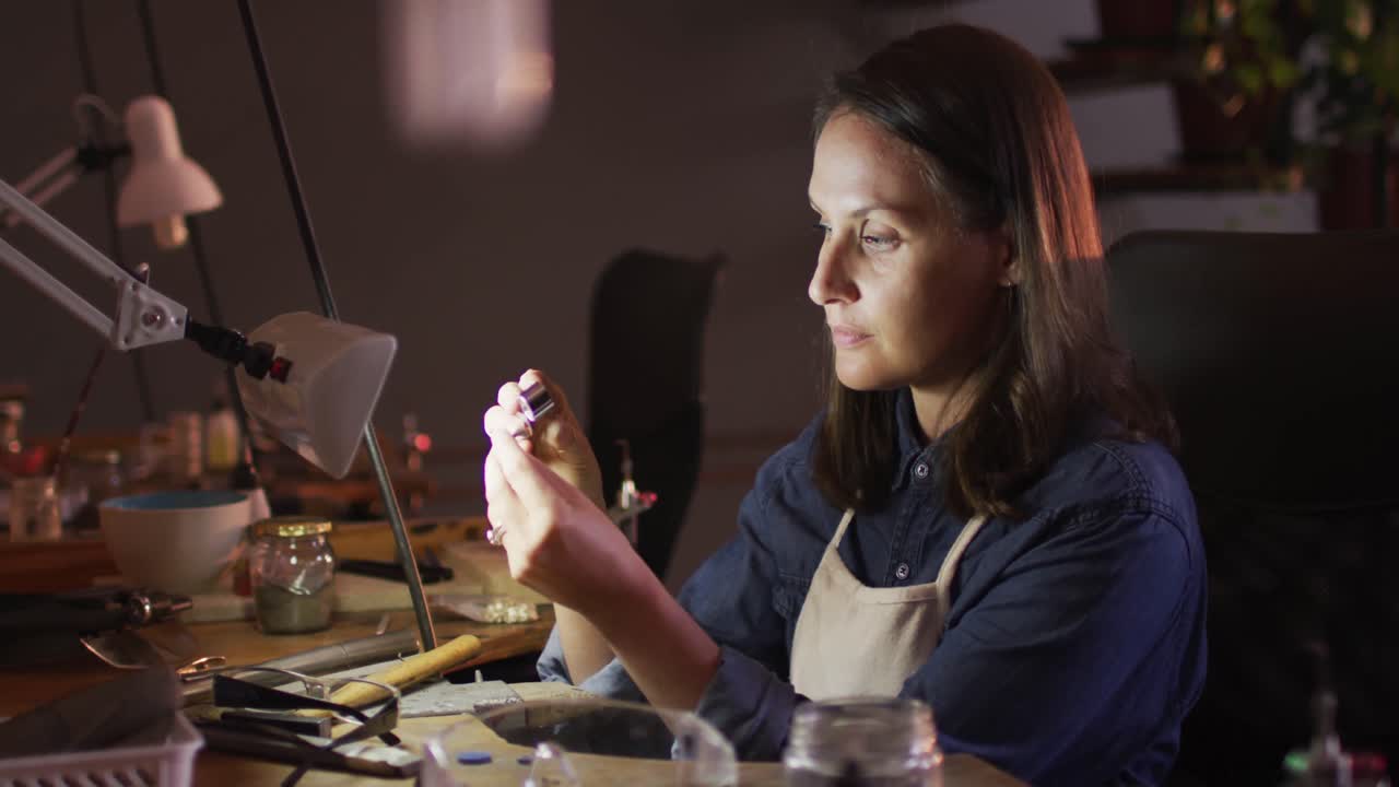 Profile of focused caucasian female jeweller sitting at desk, making jewelry in workshop