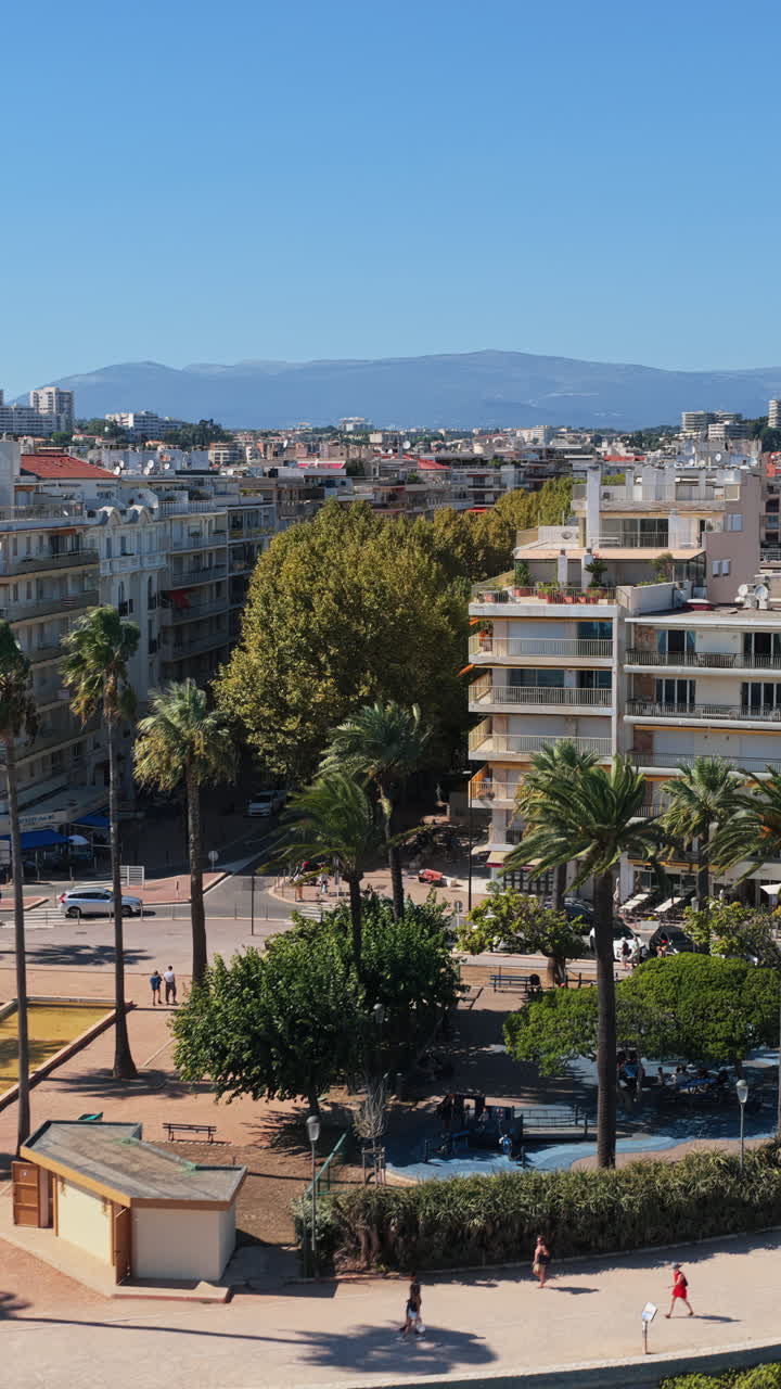 Vista aerea da drone del lungomare di Antibes, fiancheggiato da palme e appartamenti moderni, con il Mar Mediterraneo e le montagne in lontananza sullo sfondo. Verticale