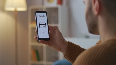 Mann-Kauft-Online,-Indem-Er-Auf-Den-Touchscreen-Seines-Smartphones-Tippt-Und-Mit-Karte-Bezahlt.-Grafiken-Auf-Dem-Display,-Ansicht-Durch-Die-Schulter-Des-Benutzers,-Der-Zu-Hause-Sitzt