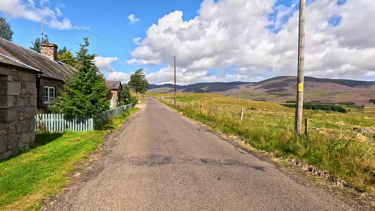 Un veicolo percorre una stretta strada di campagna attraverso la panoramica Clova, Angus, Scozia, passando case in pietra, campi e colline sotto la luce intensa del giorno con un movimento costante della telecamera in avanti.