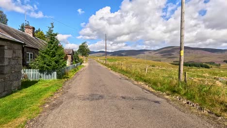 a vehicle travels a narrow country road through scenic clova, angus, scotland, passing stone houses, fields, and hills under bright daylight with steady forward camera movement