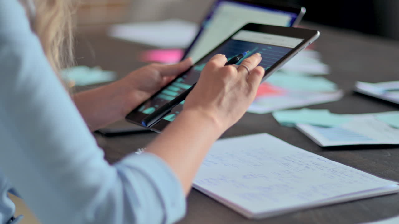 Woman in blue blazer analysing a graph on a tablet and writing in a notebook at a desk in an office