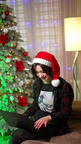 woman wearing santa cap and checkered shirt rejoices looking at laptop. happy news online. christmas tree at backdrop. vertical video.