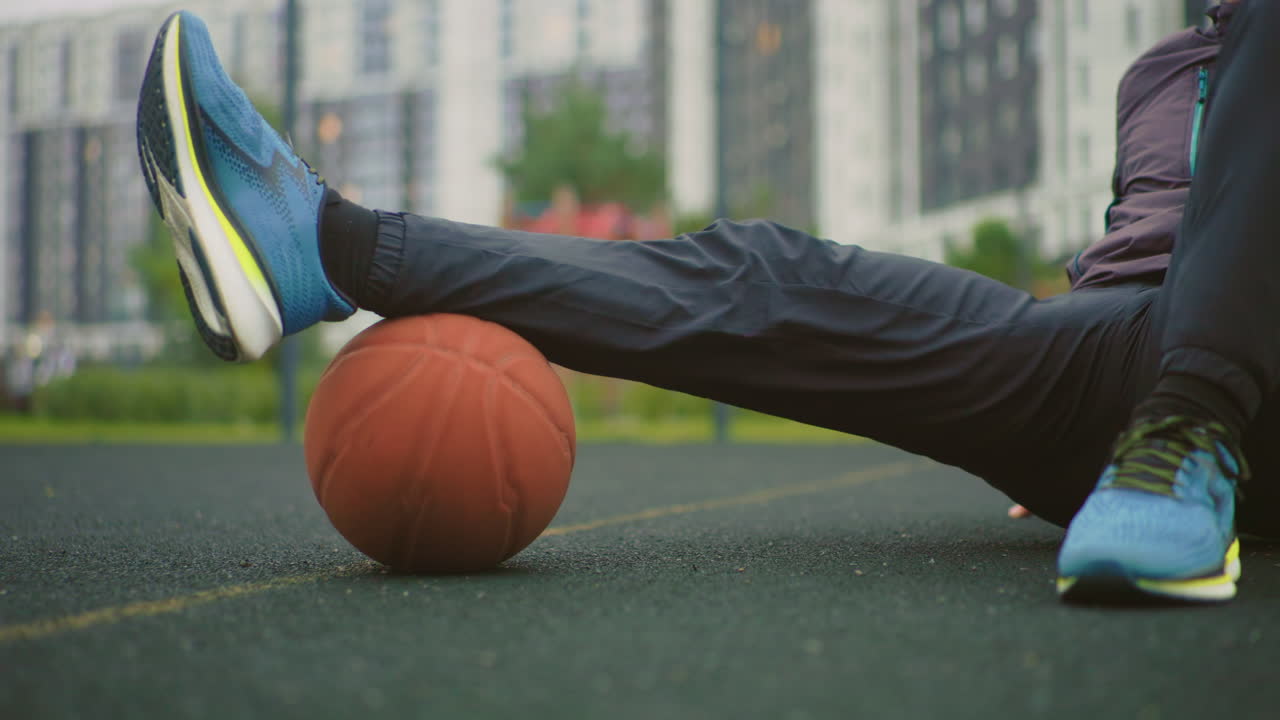 athlete on court with basketball balancing while sitting on asphalt near apartment blocks wearing blue sneakers and dark joggers practicing ball control and leg coordination lowangle cinematic frame