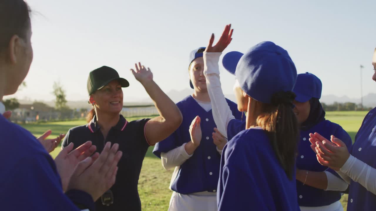 un grupo diverso de jugadoras de béisbol y entrenadoras en el campo, aplaudiendo y dando los cinco.