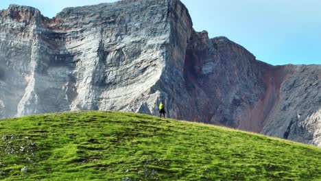 Enge-162-mm-Drohnenaufnahme-Einer-Wanderin,-Die-Auf-Einem-Hügel-Läuft,-Mit-Einer-Riesigen-Vertikalen-Felswand-Eines-Berges-Hinter-Ihr-In-Den-Dolomiten