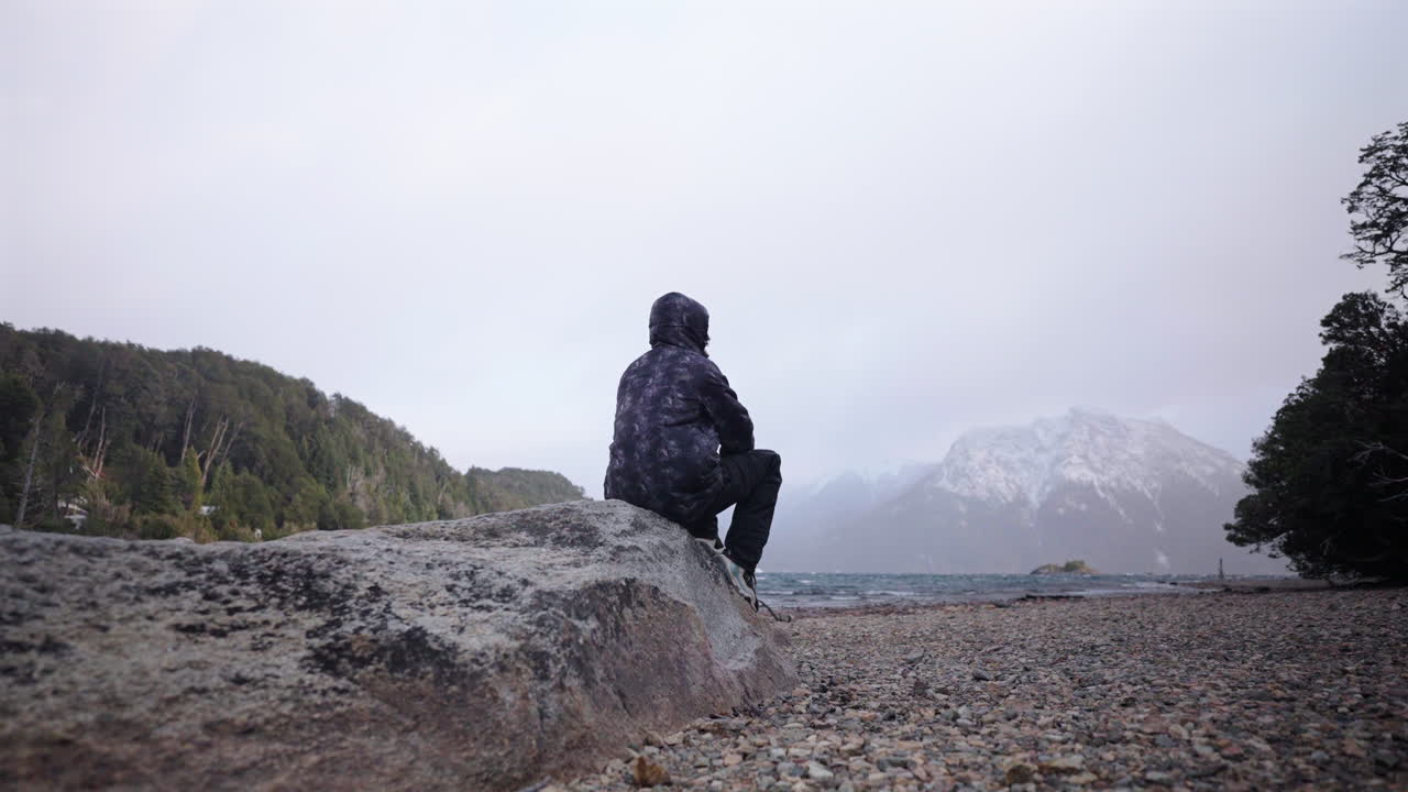Traveler finds peace while sitting near cold lake with snow covered mountains beyond