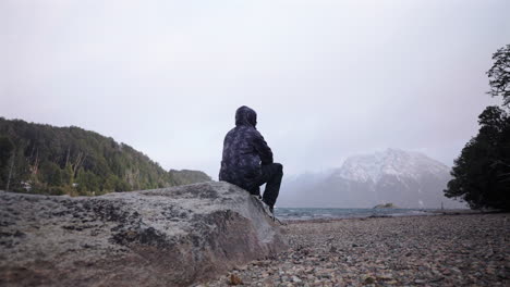 traveler finds peace while sitting near cold lake with snow covered mountains beyond
